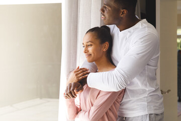 Happy diverse couple embracing and looking out of window smiling at home