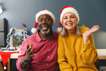 Happy diverse senior couple in santa hats for christmas video call, smiling and gesturing to camera