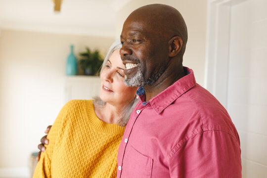 Happy Diverse Senior Couple Embracing, Looking Away And Smiling At Home