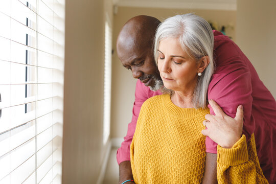 Thoughtful, sad diverse senior couple embracing and looking down at home