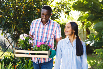 Happy diverse couple carrying flowers for planting walking in garden smiling