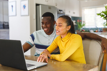 Happy diverse couple making video call on laptop smiling and talking to screen in kitchen