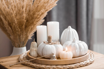 Fototapeta na wymiar Still-life. Knitted pumpkin, pumpkin-shaped candles and white ceramic pumpkins on a wooden tray on a coffee table in the home interior of the living room. Cozy autumn concept.