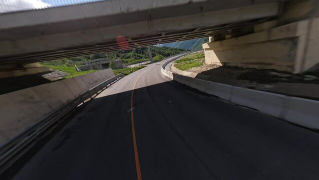 FPV sport drone dynamic shot diving over empty serpentine asphalt road mountain alpine resort sunny landscape aerial view. Speed flight in bridge tunnel picturesque suburb city landmark infrastructure