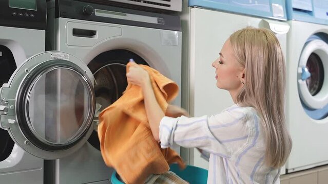 A young pretty woman loads dirty clothes into a household washing machine, which is located in a public laundry