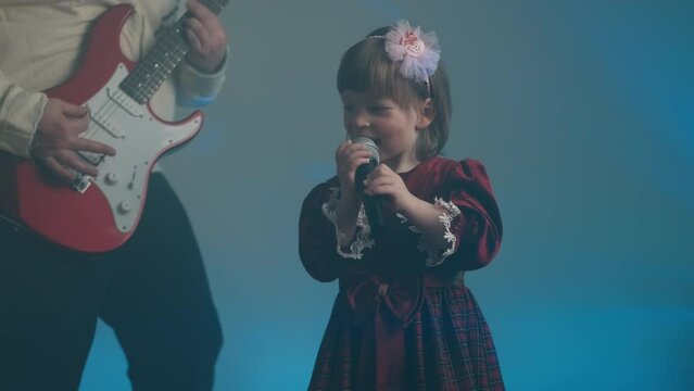 Little girl on stage in vintage dress, she sings into microphone and dances, her father plays an electric guitar. Color music is shining and smoke is billowing. Family time, creativity and hobbies