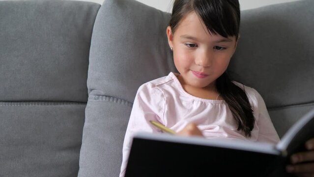 Little girl is sitting on sofa and writing the essay letter in notebook at home. Online learning, homeschooling, study from home. Kid is thinking what to write, doing homework concept. 