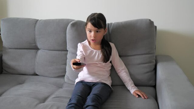 Cute little kid sitting on couch watches TV with interest, holding tv remote in hand. Cute little girl watching cartoon movie. Leisure and entertainment, happy childhood concept. 