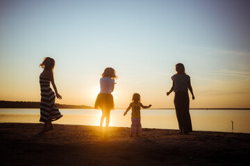 Silhouettes of children and their mothers jumping and having fun on the beach in sunset light. Good mood and pastime among the younger and older generation.