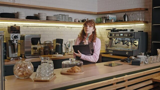 Pensive female barista makes notes in clipboard at workplace