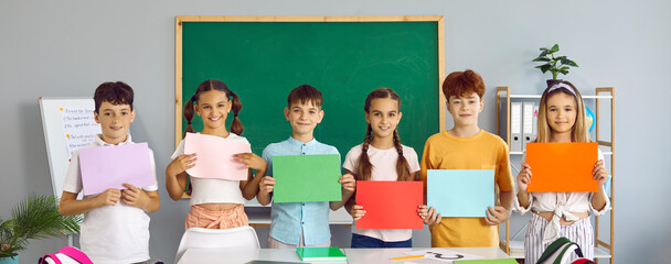 Group of happy students standing in row in classroom and holding different colorful mockup banners. Team of joyful primary junior school children study new words and show text copy space paper posters