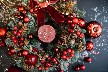 Obraz na płótnie Canvas Beautiful festive hand made wreath decorated with fresh nobilis spruce, red ornaments, berries, pine cones, candle, acorns, fake snow, decorative apples, glitter with fireflies on the background