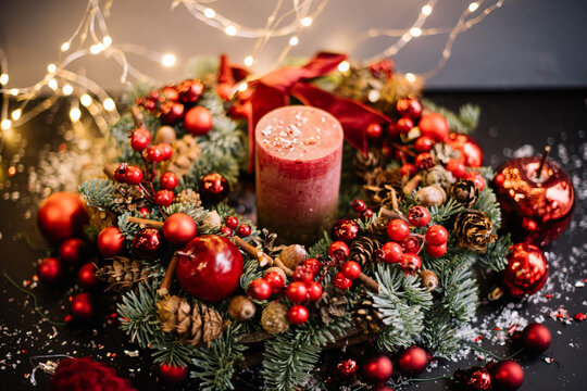 Beautiful festive hand made wreath decorated with fresh nobilis spruce, red ornaments, berries, pine cones, candle, acorns, fake snow, decorative apples, glitter with fireflies on the background - Powered by Adobe