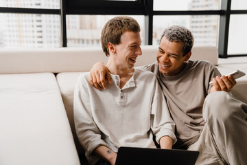 Happy gay couple using cellphone and laptop while resting at home