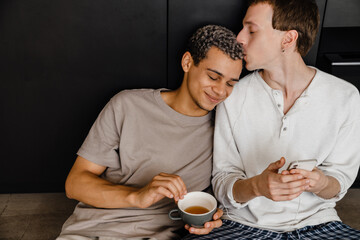 Happy young gay couple using cellphone and drinking coffee at home