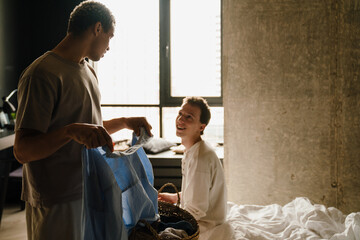Happy multiracial gay couple talking and folding clothes in bedroom