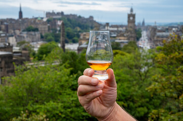 Hand holding glass of single malt scotch whisky and view from Calton hill to park and old parts of Edinburgh city in rainy day, Scotland, UK