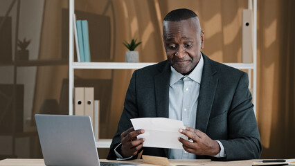 Fototapeta na wymiar African old man senior mature male in office opening paper envelope reading letter with good news. Happy elder retired businessman received investment notification banking loan approval salary bonus