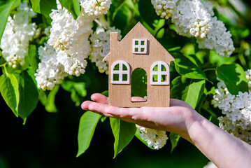 The girl holds the house symbol against the background of blossoming white lilac