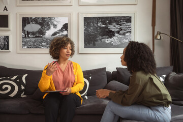 A female psychotherapist talks to the teenage African-American girl who sits on a couch at her office. The girl listens carefully to what the therapist tells her