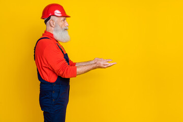 Photo of positive good mood senior guy dressed uniform overall red hardhat holding arms empty space isolated yellow color background