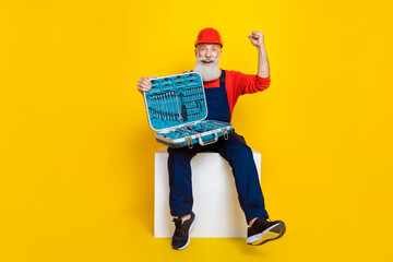 Photo of funny lucky age man wear overall uniform red hard hat rising fist open toolset isolated yellow color background