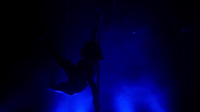 Slim woman performing poledance tricks sensually in spotlights. Sexy lady making moves upside down in night club. Young blonde dancer doing twine on pylon illuminated soft nightclub backlight.