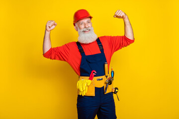 Photo of cool cheerful age man wear overall uniform red hard hat showing biceps rising fists isolated yellow color background