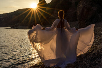 A mysterious female silhouette with long braids stands on the sea beach with mountain views, Sunset...