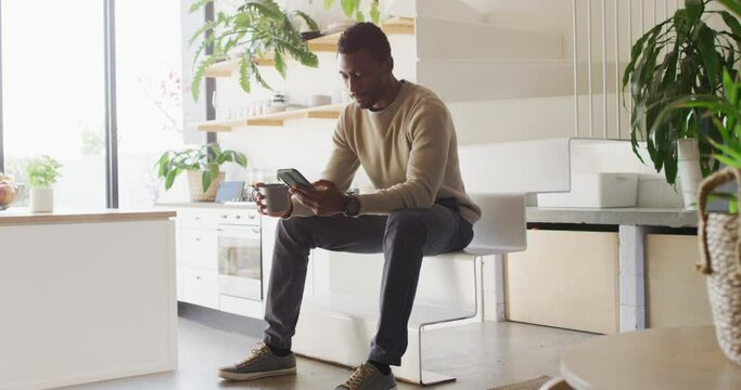 Happy african american man sitting on stairs in kitchen, drinking coffee and using smartphone