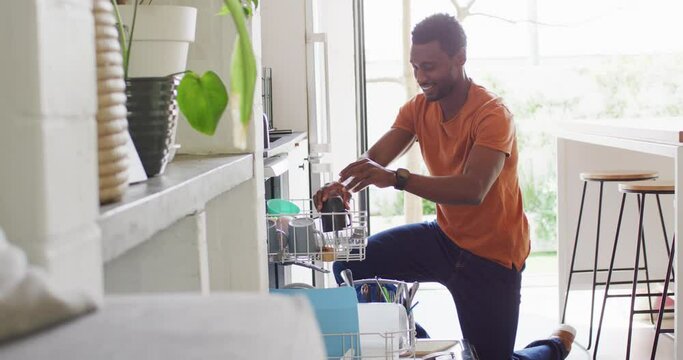 Happy african american man standing in kitchen and loading dishwasher