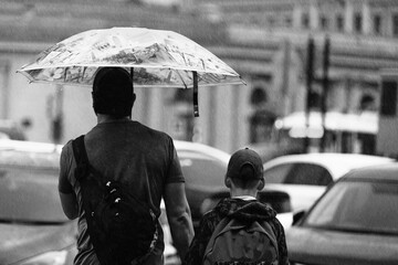 Father and son walk among cars in the rain under an umbrella, black and white photo.