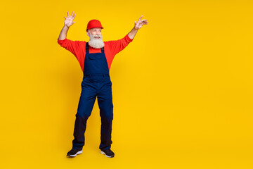 Photo of funny positive age man workwear overall red hard hat dancing empty space isolated yellow...