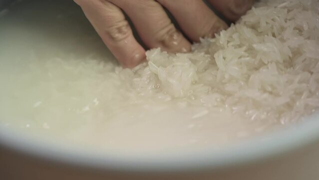 Close up of hand washing white Thai jasmine long grain rice with water in a rice cooker bowl in slow motion.