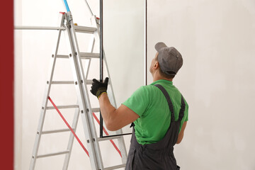 Worker holding double glazing near ladder indoors. Window installation - obrazy, fototapety, plakaty