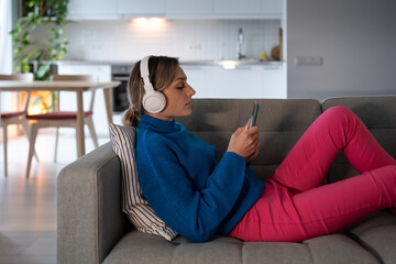 Obraz premium Young woman lying on comfortable sofa rests from hard work in cozy apartment. Female wearing white headphones searches music to listen and scrolls news in Internet social media on smartphone