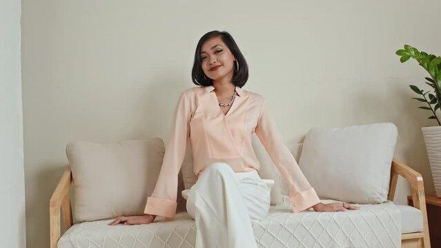 Pretty Asian model in elegant pastel outfit sitting on sofa in studio and posing for camera during fashion photoshoot