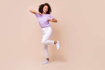 Fototapeta na wymiar Full size photo of adorable positive woman wear striped t-shirt white trousers sneakers dancing isolated on beige color background