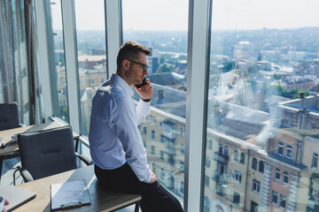 A young businessman with a phone in his hands in glasses and a white shirt, he is in a modern office with large windows. A man in classic trousers and a white shirt. Young attractive businessman