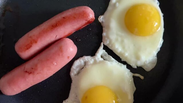 Cooking fried eggs with sausages in frying pan in home kitchen. Traditional American or English breakfast. Close-up.
