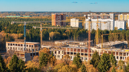 Fototapeta na wymiar Construction site panorama. Construction business of real estate and residential buildings. Build in forest area in Troitsk city. Cranes against woods trees and city in autumn sunny day