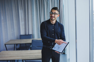 A Caucasian business man in glasses and a black shirt sits in the office and looks at sales growth charts. A young successful businessman in classic clothes stands by the window in the office