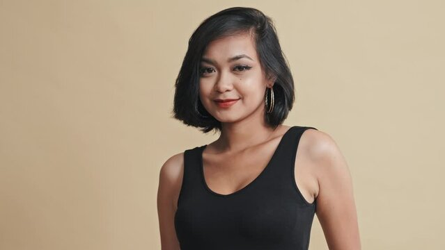 Tilt up shot of Asian woman in stylish outfit posing on camera against beige background during fashion lookbook photoshoot in studio