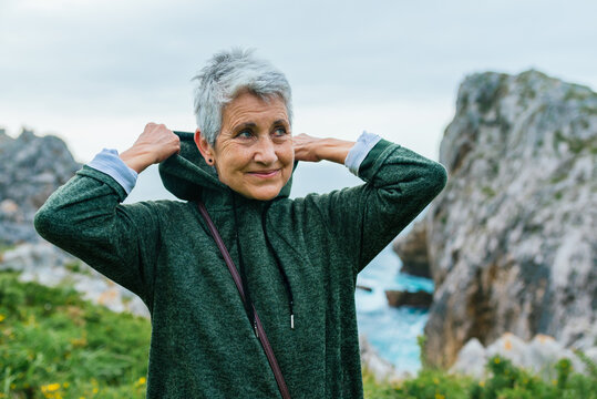 Older Woman With Gray Hair, Wearing Sportswear, Pulling Up The Hood Of Her Sweatshirt As She Jogs Along A Coastal Path. Sports Activities In The Elderly.