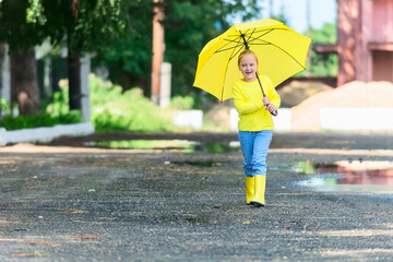 a schoolgirl girl of seven years old in yellow clothes and with a bright yellow umbrella in her hands, in rubber waterproof boots in the warm season walks through the puddles after the rain