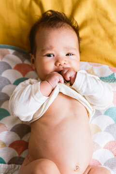 Close Up Of Cute Latino Baby Girl Eating Her T Shirt While Looking In Camera. Waking Up In Bed. Healthy Hispanic Baby With Warm Natural Light