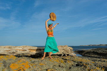 A woman in boho style clothes with a shamanic ritual tambourine on the rocks on the seashore on a clear sunny day. The concept of freedom of mind and body in nature
