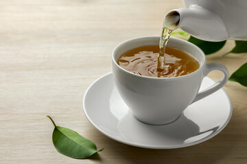 Pouring green tea into white cup with saucer and leaves on wooden table, closeup. Space for text