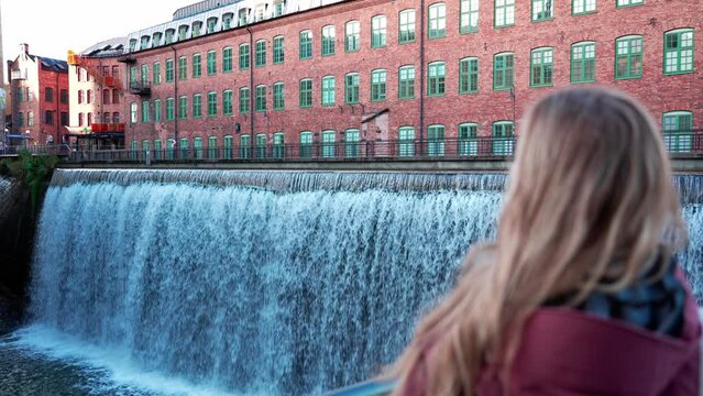 Blonde girl stands in front of a large waterfall in central Norrköping, Motala Ström river. Tourism in Sweden, Scandinavian culture and civil engineering. Industrial city in southern Sweden. Travel, m