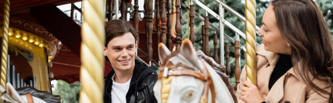cheerful young man looking at girlfriend riding carousel horse in amusement park, banner.